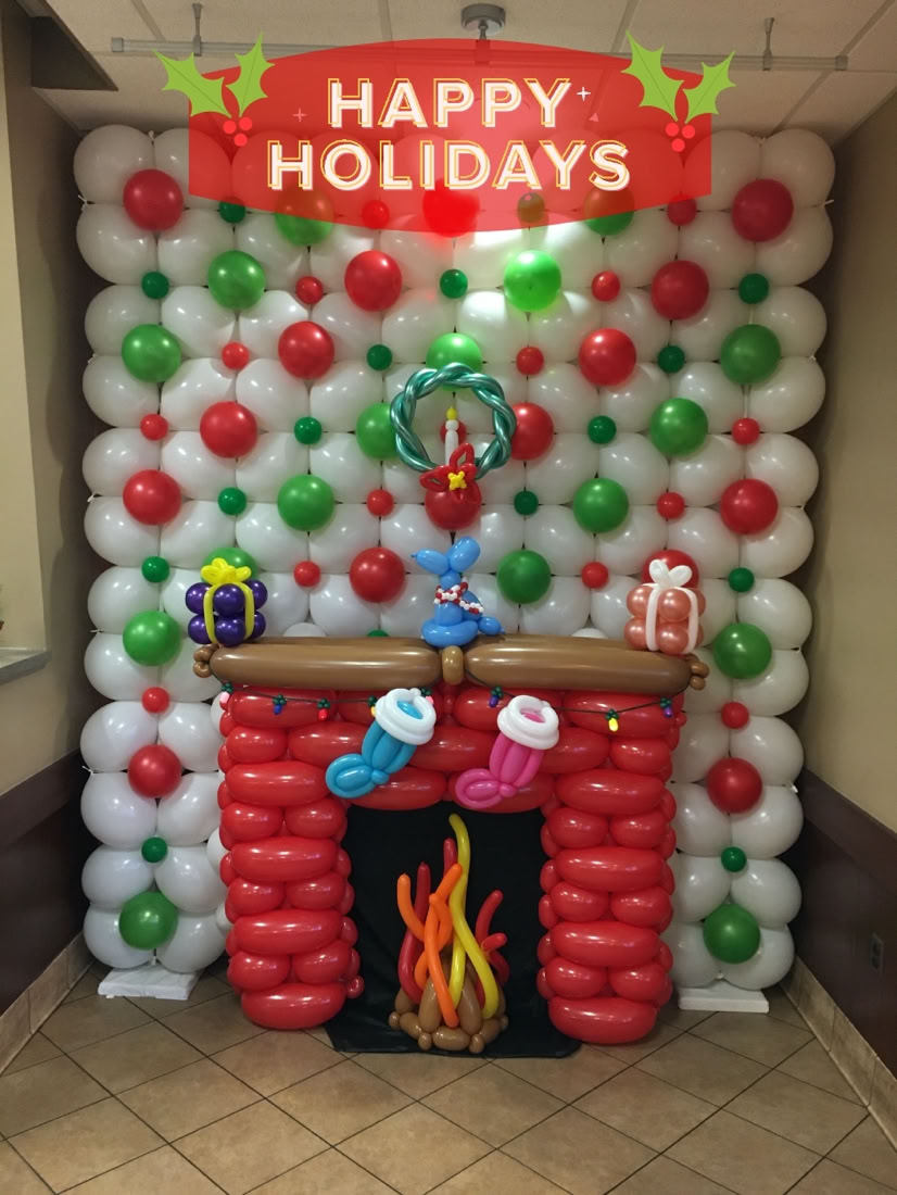 This image shows a balloon sculpture of a Christmas-themed fireplace scene set up indoors. * The entire wall is covered in white balloons with evenly spaced red and green balloon accents, creating a festive, holiday-inspired backdrop. * In the center is a fireplace, made entirely of balloons, but appearing to be built of red brick with a brown mantle. * Inside the fireplace is a colorful flame, crafted with twisted balloons in orange, yellow, and red, giving the illusion of a roaring fire. * Hanging from the mantle are two Christmas stockings also made entirely of balloons, one blue and one pink, with white tops. * On top of the mantle are three balloon gift boxes, each topped with a bow—colored in purple, red, and white. * Above the fireplace, there is a green balloon wreath with a red bow, suspended from the backdrop. * The floor is tiled, and the scene is set in a corner of a room, for a Christmas party or holiday event. This delightful balloon art setup captures the warmth and fun of the holiday season with a whimsical, creative touch.
