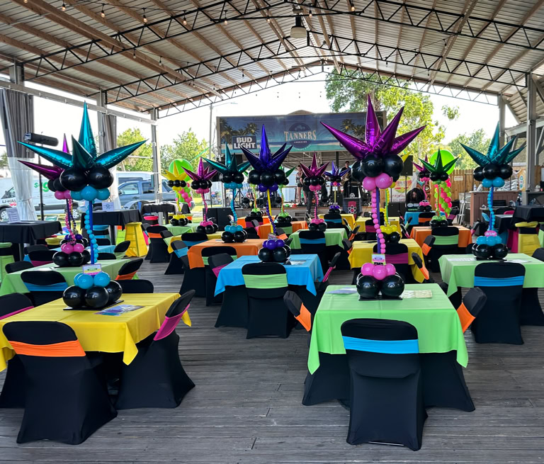 This image shows a festive party or event setup under a large, open-air pavilion with a high metal roof. The area is decorated with brightly colored tables and balloon centerpieces, creating a vibrant and lively atmosphere. Each square table is draped with a brightly colored tablecloth—yellow, green, orange, or blue—and surrounded by chairs covered in black fabric with colorful sashes in matching or contrasting colors. At the center of each table is a large and whimsical balloon arrangement. These centerpieces are made of clusters of shiny balloons in various colors (like teal, purple, pink, and blue) and shaped to resemble abstract starbursts or fireworks. The balloon sculptures rise from the center of the tables and feature spiral stems made from smaller balloons, adding a sense of movement and fun. In the background, there is a small stage area with musical equipment. The ceiling of the pavilion is lit with purple and blue lighting, adding to the celebratory vibe. Overall, the scene is colorful, cheerful, and clearly designed for a lively and fun event.