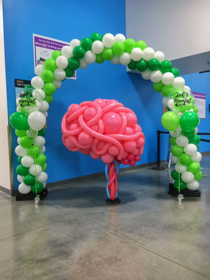 This image captures a lively indoor celebration filled with a large crowd of people. The ceiling is draped with white fabric and lit with colorful lights—purple, pink, and orange hues. Dozens of oversized balloons in vibrant colors (red, orange, purple, green, and blue) are floating and bouncing above the excited crowd. Many people have their hands raised, reaching for the balloons, while others are taking photos. The atmosphere is festive and energetic, resembling a balloon drop party or grand finale event.