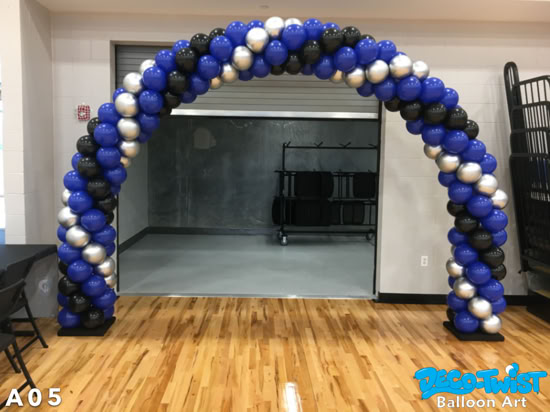 A balloon arch made of blue, black, and silver balloons stands indoors on a gymnasium floor, creating a bold and festive entryway.