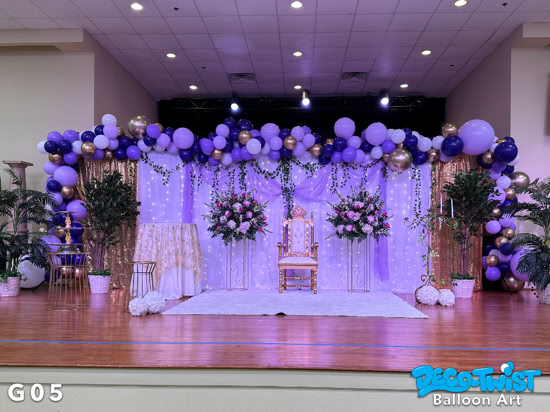 A balloon garland in shades of purple, lavender, white, and gold decorates the top of an elegant stage backdrop. The setup features a gold chair, floral arrangements, greenery, and draped lights for a royal, festive look.