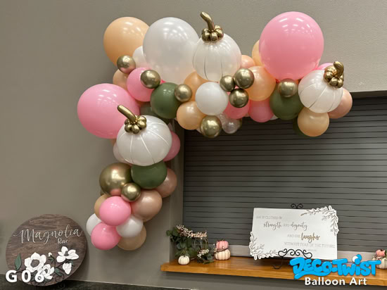A balloon garland in soft fall colors—peach, pink, white, green, and gold—decorates a wall corner. Some balloons are shaped like pumpkins with gold stems, adding an autumn harvest theme.