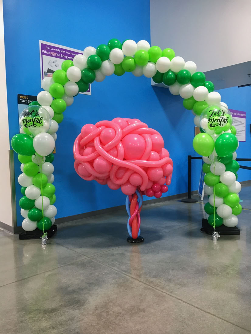 This image shows a balloon display in a public indoor space, for an event promoting mental health awareness. * The main feature is a large balloon sculpture of a brain, made with various shades of pink balloons twisted and intertwined to mimic the appearance of brain folds. The brain is supported by a base made of twisted red and blue balloons representing the brain stem. * Surrounding the brain is a balloon archway made of white and multiple shades of green balloons arranged in a spiral pattern. The arch is supported by a sturdy black bases on either side. * On both vertical sides of the arch are floating balloon bouquets crowned with a clear bubble balloon featuring custom black vinyl text that reads “Let’s Talk About Mental Health”. The balloon brain and arch create a vibrant, engaging visual centerpiece likely intended to draw attention and promote a message related to mental health.