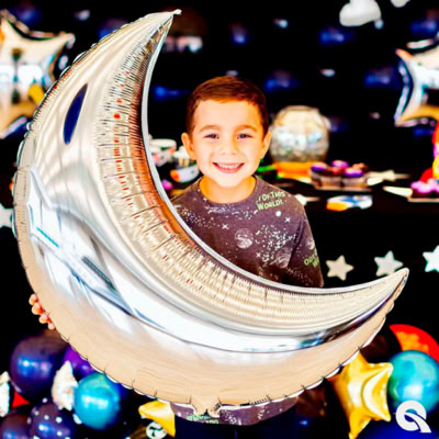 A young boy is smiling while holding a large silver balloon shaped like a crescent moon. He is wearing a space-themed shirt with stars and planets on it. The background is decorated with balloons and space-themed items, suggesting a space-themed party.