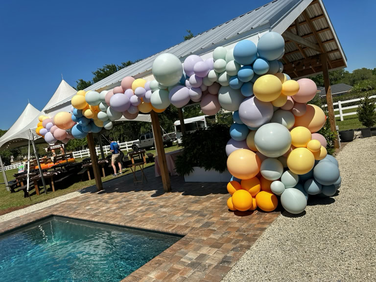 This image captures an organic balloon garland installation at an outdoor event. * The focal point is a large, pastel-colored balloon garland attached to the side and roofline of a wooden pavilion. The balloons vary in size and include soft colors such as peach, lavender, baby blue, pale yellow, mint green, and light orange. The arrangement creates a cascading effect that covers the right corner post and continues across the top of the structure. * The setting is bright and sunny, with a clear blue sky overhead, enhancing the cheerful, airy atmosphere. * In the foreground is a pool with clear, sparkling water bordered by brick pavers. * In the background, there are white event tents and picnic tables. * The overall scene is calm and inviting, blending natural elements with decorative flair — perfect for a spring or summer birthday party, shower, or outdoor brunch. This display shows how balloons can add a soft, elegant, and playful touch to upscale outdoor gatherings.