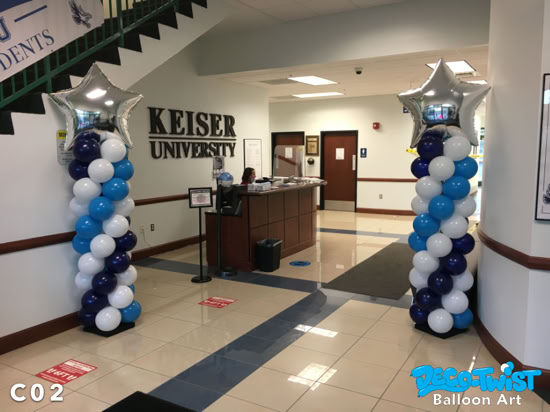 Two balloon columns stand inside a university lobby, each topped with a large silver star balloon. The columns are spiraled with navy blue, light blue, and white balloons, creating a bold and festive display at the entrance area.