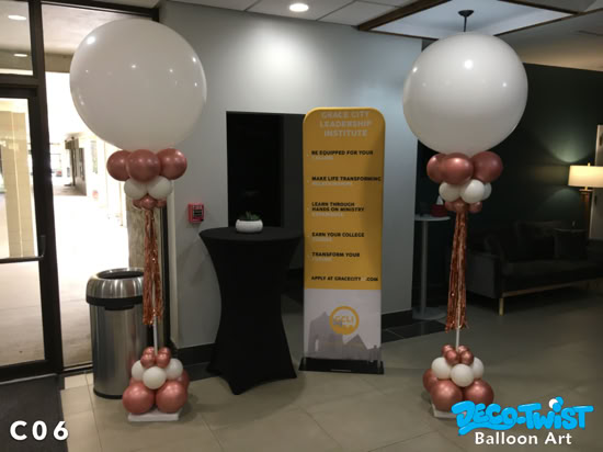 Two elegant balloon columns stand indoors, each topped with a very large white balloon. Below the giant balloons are clusters of smaller rose-gold and white balloons, with metallic tassels hanging down for decoration. The bases also feature balloon clusters in matching colors. A tall yellow banner in the background promotes a leadership institute.