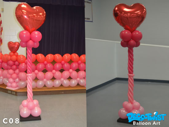 This image shows two tall balloon columns designed with a Valentine’s theme. Each column features a large shiny red heart balloon on top, supported by clusters of smaller pink balloons. The column shafts are made of long pink balloons twisted in a spiral braid, with a base of round balloons for support. The left side shows the column placed in front of a stage decorated with rows of pink and red balloons, while the right side shows the column standing alone in a plain corner. The design is festive, romantic, and eye-catching, often used for parties, dances, or Valentine’s events.