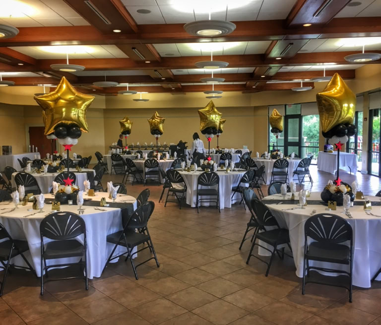 This image showcases a sophisticated indoor party or banquet setup with a “Hollywood” or “Awards Night” theme. The atmosphere is elegant and celebratory, with round dining tables dressed in white tablecloths and stylish black chairs arranged neatly around them. Each table features a striking balloon centerpiece. These centerpieces are made up of a large, shiny gold foil balloon shaped like a star, sitting above clusters of smaller black and white balloons. The base of the balloon cluster is decorated with additional elements, including small red star cutouts and shiny gold accents, adding to the glitzy feel. A tall black centerpiece frame holds the balloon clusters aloft, making them stand tall and prominent over each table. A shimmering black runner runs across the center of the table, adding contrast to the white linens. Overall, the scene is an upscale event perfect for a gala, awards ceremony, graduation party, or formal celebration of any kind.