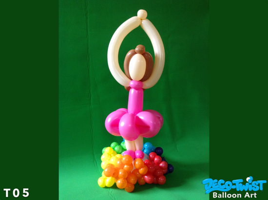 This image shows a balloon sculpture of a ballerina. The ballerina is made from twisting balloons, with beige balloons forming her arms raised overhead in a dance pose, and brown balloons for her hair. Her tutu is created from bright pink balloons, and she stands on a colorful base made of small balloons in rainbow colors. The background is a plain green backdrop, highlighting the playful and artistic balloon centerpiece.