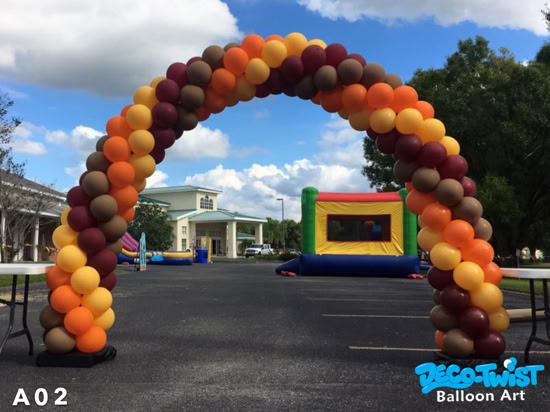 A large balloon arch made of brown, orange, yellow, and maroon balloons stands outdoors. It frames the view of a bounce house and event area in the background.