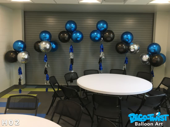 This image shows several latex-free balloon bouquets arranged along a wall. Each bouquet is made of shiny helium balloons in metallic blue, black, and silver, with decorative tassel tails hanging beneath them. The balloons are placed behind round white tables with black chairs set up for seating, creating a festive and modern event setup.