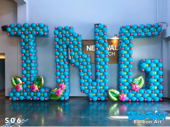 A giant balloon display spells out the letters “ING” in bright blue balloons with small pink accents. Each letter is approx. 12' tall. The base is decorated with green leaf-shaped balloons and pink flower balloons.