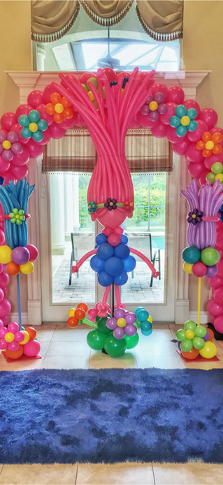 This image depicts a colorful and whimsical balloon decoration setup inside a spacious room with elegant furnishings. The focal point is a large arch made entirely of balloons, creating a playful and welcoming entrance. The arch is composed mainly of bright pink balloons, accented with balloon flowers in vibrant colors like turquoise, orange, yellow, purple, and green. These balloon flowers are spaced evenly along the arch and add a cheerful, garden-like theme. At the center of the arch is a balloon figure designed to look like a character, likely inspired by Poppy from the movie Trolls.  To the left and right of the arch, there are two additional balloon structures with similar color schemes and flower designs, enhancing the symmetry and theme. A dark blue plush rug lies on the tile floor in front of the setup. Overall, the scene is festive and ideal for a child’s party or a themed event, evoking joy and fantasy.