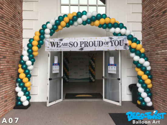 A large balloon arch made of green, gold, and white balloons frames the entrance of a building. A banner hanging across the arch reads, “We are so proud of you,” celebrating a graduation.