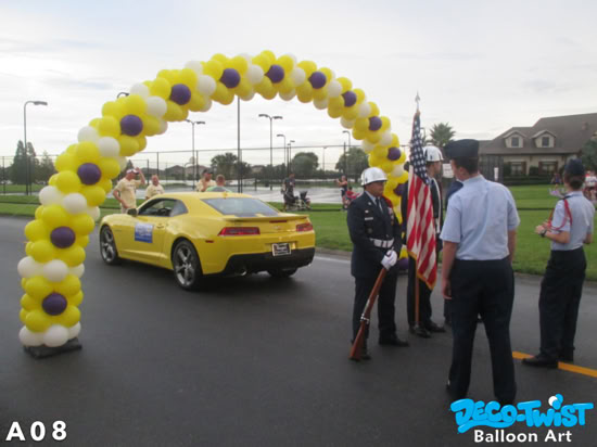 A large balloon arch made of yellow, white, and purple balloons stands over a street. Beneath it, a yellow sports car and a color guard holding the American flag are participating in a parade or ceremony.