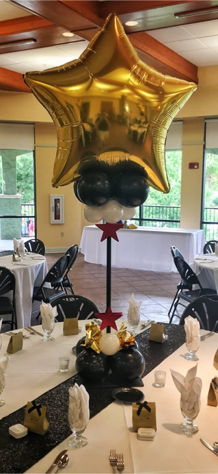 This image showcases a sophisticated indoor party or banquet setup with a “Hollywood” or “Awards Night” theme. The atmosphere is elegant and celebratory, with round dining tables dressed in white tablecloths and stylish black chairs arranged neatly around them. Each table features a striking balloon centerpiece. These centerpieces are made up of a large, shiny gold foil balloon shaped like a star, sitting above clusters of smaller black and white balloons. The base of the balloon cluster is decorated with additional elements, including small red star cutouts and shiny gold accents, adding to the glitzy feel. A tall black centerpiece frame holds the balloon clusters aloft, making them stand tall and prominent over each table. A shimmering black runner runs across the center of the table, adding contrast to the white linens. Overall, the scene is an upscale event perfect for a gala, awards ceremony, graduation party, or formal celebration of any kind.