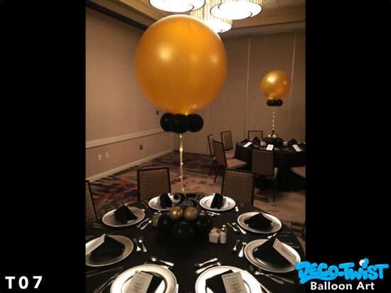 This image shows an elegant event setup with round dining tables. Each table is decorated with a large gold balloon centerpiece anchored by smaller black balloons. The tables are set with black napkins, silver chargers, and menus, creating a classy black-and-gold theme. The room has soft lighting, enhancing the formal and celebratory atmosphere.