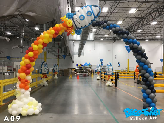 A balloon arch designed to look like a rocket launch. One side of the arch is made of red, orange, and yellow balloons representing fire and smoke, while the other side is blue, gray, and black balloons leading up to a rocket-shaped balloon at the top.