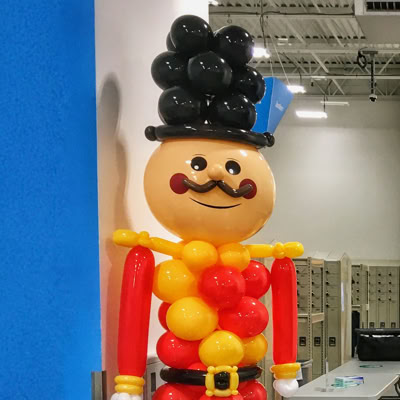 The image shows a large balloon sculpture of a cheerful toy soldier. It has a round face with a mustache, rosy cheeks, and a big smile. The body is made of red and yellow balloons, with black boots and a tall black balloon hat. The figure is set up indoors.