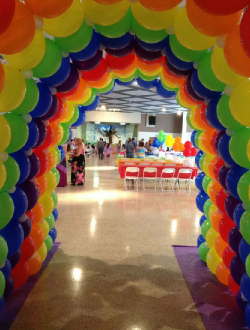 This image shows a vibrant rainbow-colored balloon tunnel leading into a festive event space. * The tunnel is made entirely of balloons in rows of rainbow colors: red, orange, yellow, green, blue, and purple. * The tunnel forms a colorful passageway that guests walk through to enter the event space. * At the end of the tunnel, you can see an event space with tables covered in bright cloths and balloon centerpieces, and a few people mingling. * The floor is a shiny, speckled surface, and there’s a purple carpet under the tunnel entrance, adding a special touch to the entry. * The overall scene gives off a cheerful and welcoming vibe.