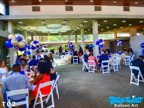 This image shows a lively banquet hall filled with round tables covered in light-colored tablecloths and surrounded by white chairs. Each table features a balloon centerpiece made of gold, royal blue, and clear confetti-filled balloons, arranged in clusters. Guests, dressed in formal attire, are seated and enjoying food and conversation at this wedding reception.