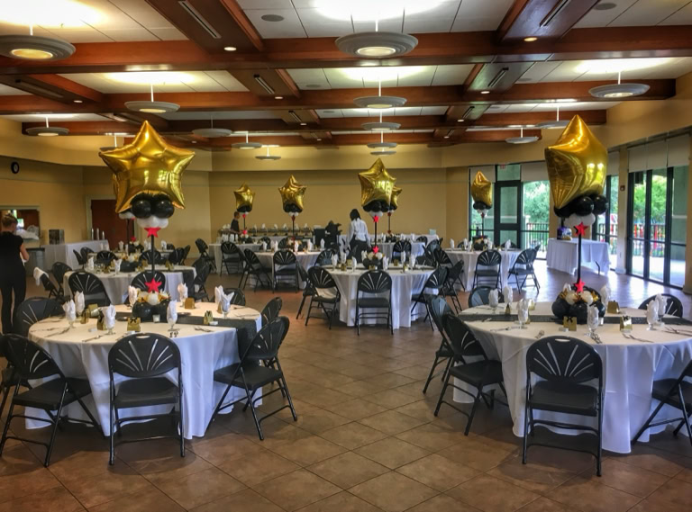 This image showcases a sophisticated indoor party or banquet setup with a “Hollywood” or “Awards Night” theme. The atmosphere is elegant and celebratory, with round dining tables dressed in white tablecloths and stylish black chairs arranged neatly around them. Each table features a striking balloon centerpiece. These centerpieces are made up of a large, shiny gold foil balloon shaped like a star, sitting above clusters of smaller black and white balloons. The base of the balloon cluster is decorated with additional elements, including small red star cutouts and shiny gold accents, adding to the glitzy feel. A tall black centerpiece frame holds the balloon clusters aloft, making them stand tall and prominent over each table. A shimmering black runner runs across the center of the table, adding contrast to the white linens. Overall, the scene is an upscale event perfect for a gala, awards ceremony, graduation party, or formal celebration of any kind.