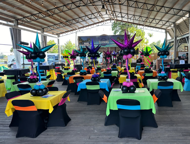 This image shows a festive party or event setup under a large, open-air pavilion with a high metal roof. The area is decorated with brightly colored tables and balloon centerpieces, creating a vibrant and lively atmosphere. Each square table is draped with a brightly colored tablecloth—yellow, green, orange, or blue—and surrounded by chairs covered in black fabric with colorful sashes in matching or contrasting colors. At the center of each table is a large and whimsical balloon arrangement. These centerpieces are made of clusters of shiny balloons in various colors (like teal, purple, pink, and blue) and shaped to resemble abstract starbursts or fireworks. The balloon sculptures rise from the center of the tables and feature spiral stems made from smaller balloons, adding a sense of movement and fun. In the background, there is a small stage area with musical equipment. The ceiling of the pavilion is lit with purple and blue lighting, adding to the celebratory vibe. Overall, the scene is colorful, cheerful, and clearly designed for a lively and fun event.