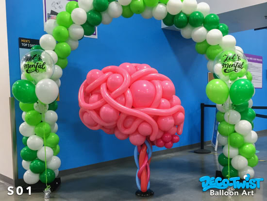 A large balloon sculpture shaped like a pink brain stands under a green and white balloon arch. The setup is festive and colorful, with balloons arranged to promote a mental health awareness event.