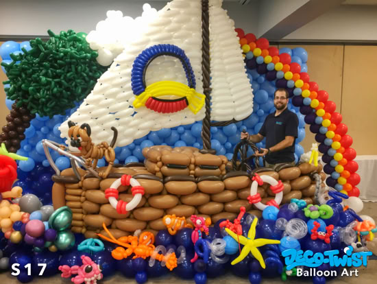 A large balloon sculpture depicts a sailboat at sea. The boat is made of brown balloons with life preservers, a white sail, a rainbow arch, and colorful ocean animals below. A man stands inside the balloon boat holding the wheel.