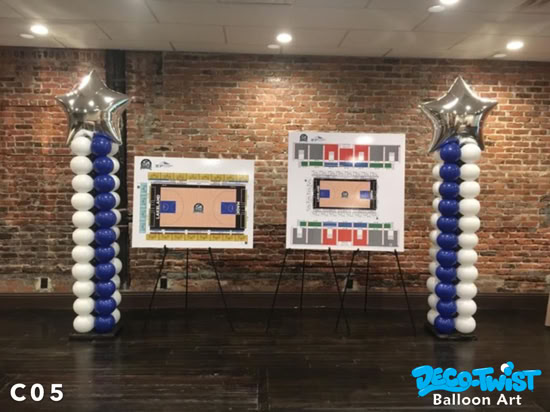 Two tall balloon columns stand in front of a brick wall, each made of alternating blue and white balloons stacked neatly. At the top of each column is a large silver star-shaped balloon. Between the columns are two easels displaying large poster boards with sports court and seating layout diagrams.