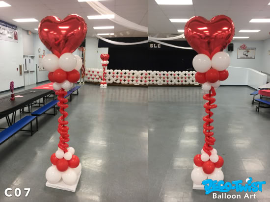 Two festive balloon columns decorated in red and white stand in a gym-like space. Each column is topped with a large red foil heart balloon, followed by clusters of white and red balloons. The main stems of the columns are made with curly red balloons spiraling downward, ending in balloon bases of red and white. They create a romantic, Valentine’s Day–themed atmosphere.