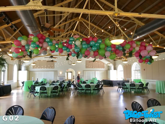 A large balloon garland made of pink, green, teal, and white balloons is suspended from the ceiling of the Magnolia Building in Lakeland, Florida adding festive color above round tables set for guests.