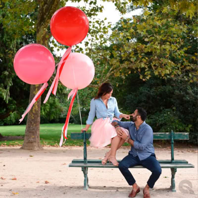 A couple is sitting on a green bench in a park surrounded by trees. The woman, dressed in a light blue top and a soft pink tulle skirt, is perched on the backrest of the bench holding large helium balloons in shades of red and pink with flowing ribbons. The man, wearing a blue shirt and dark pants, is sitting on the bench, looking up at her affectionately while gently touching her knee. The atmosphere is romantic and serene, with soft sunlight filtering through the trees.