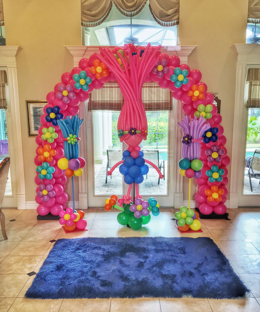 This image depicts a colorful and whimsical balloon decoration setup inside a spacious room with elegant furnishings. The focal point is a large arch made entirely of balloons, creating a playful and welcoming entrance. The arch is composed mainly of bright pink balloons, accented with balloon flowers in vibrant colors like turquoise, orange, yellow, purple, and green. These balloon flowers are spaced evenly along the arch and add a cheerful, garden-like theme. At the center of the arch is a balloon figure designed to look like a character, likely inspired by Poppy from the movie Trolls.  To the left and right of the arch, there are two additional balloon structures with similar color schemes and flower designs, enhancing the symmetry and theme. A dark blue plush rug lies on the tile floor in front of the setup. Overall, the scene is festive and ideal for a child’s party or a themed event, evoking joy and fantasy.