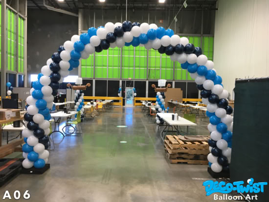 A large balloon arch made of blue, white, and dark blue balloons stands inside a warehouse, framing the walkway into an area setup for an event.