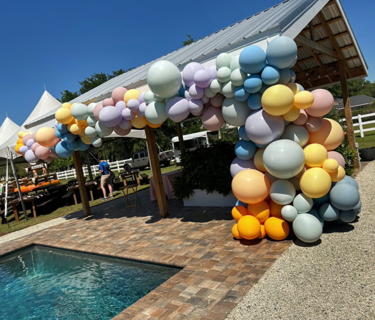 This image captures an organic balloon garland installation at an outdoor event. * The focal point is a large, pastel-colored balloon garland attached to the side and roofline of a wooden pavilion. The balloons vary in size and include soft colors such as peach, lavender, baby blue, pale yellow, mint green, and light orange. The arrangement creates a cascading effect that covers the right corner post and continues across the top of the structure. * The setting is bright and sunny, with a clear blue sky overhead, enhancing the cheerful, airy atmosphere. * In the foreground is a pool with clear, sparkling water bordered by brick pavers. * In the background, there are white event tents and picnic tables. * The overall scene is calm and inviting, blending natural elements with decorative flair — perfect for a spring or summer birthday party, shower, or outdoor brunch. This display shows how balloons can add a soft, elegant, and playful touch to upscale outdoor gatherings.