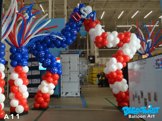 A patriotic balloon arch in red, white, and blue stands inside an Amazon Fresh warehouse. The arch forms a star shape with blue balloons decorated with white stars, and letters spelling “USA” at the top. Long red, white, and blue balloon streamers extend upward, adding a festive flair.