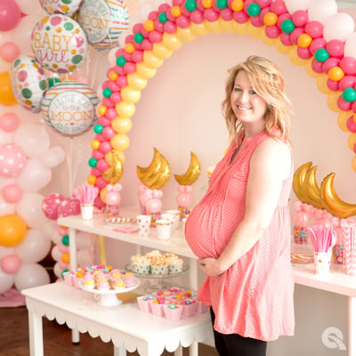 A smiling pregnant woman stands at a baby shower decorated in pink, yellow, and teal. Behind her is a colorful balloon arch, gold moon balloons, and dessert tables with sweet treats and themed decorations. The setting celebrates the upcoming arrival of a baby girl.