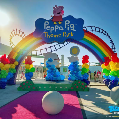 This image shows the colorful entrance to the Peppa Pig Theme Park at it's Grand Opening. A large rainbow arch with balloon decorations stretches across the entrance, topped with a playful Peppa Pig sign. The arch is adorned with bright balloon clusters in rainbow colors on both sides and fluffy balloon clouds with a sun in the center. Behind the arch, a white picket fence encloses the park, and a roller coaster track is visible in the background.