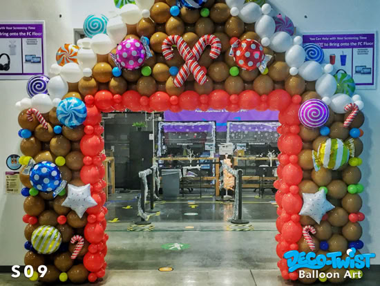 A large balloon archway is designed to look like a gingerbread house. Brown, red, and white balloons form the structure, while colorful candy-shaped balloons like lollipops, peppermints, and candy canes decorate it for a festive look.