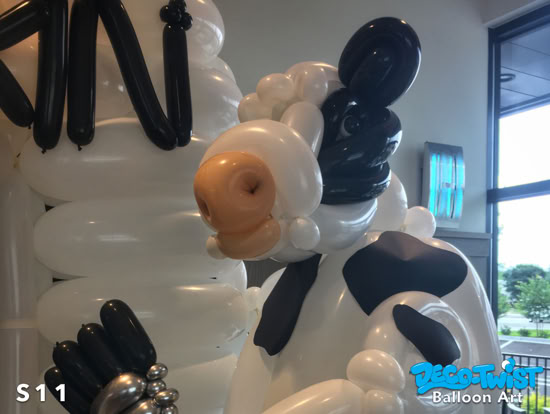 A balloon sculpture of a cow with black and white balloon patches, a light brown balloon nose, and detailed features like ears and hooves.