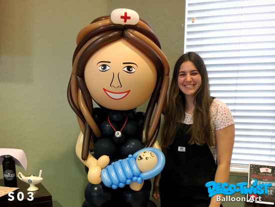 A large balloon sculpture of a smiling nurse holding a baby stands next to a young woman. The nurse figure has long brown balloon hair, a nurse’s hat with a red cross, and is dressed in black balloons with a stethoscope detail.