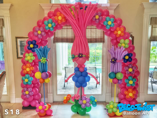 A colorful balloon sculpture creates an arch decorated with flowers and a central figure of a Troll character with tall pink balloon hair and a blue balloon dress, inspired by the Trolls movie.