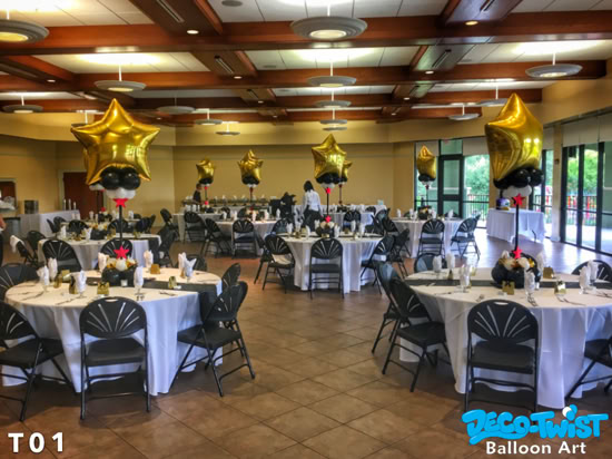 This image shows a decorated banquet hall with round tables covered in white tablecloths and black chairs arranged neatly around them. Each table has a tall centerpiece made of a large gold star-shaped balloon on top, with smaller black and white balloons beneath it. The elegant setup suggests a formal event or celebration, such as a gala, awards banquet, or corporate dinner.