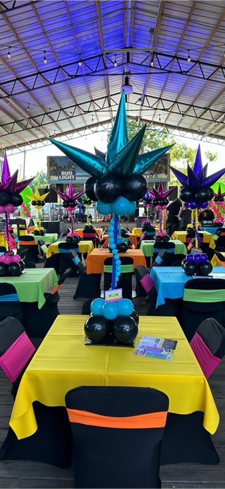This image shows a festive party or event setup under a large, open-air pavilion with a high metal roof. The area is decorated with brightly colored tables and balloon centerpieces, creating a vibrant and lively atmosphere.  Each square table is draped with a brightly colored tablecloth—yellow, green, orange, or blue—and surrounded by chairs covered in black fabric with colorful sashes in matching or contrasting colors. At the center of each table is a large and whimsical balloon arrangement. These centerpieces are made of clusters of shiny balloons in various colors (like teal, purple, pink, and blue) and shaped to resemble abstract starbursts or fireworks. The balloon sculptures rise from the center of the tables and feature spiral stems made from smaller balloons, adding a sense of movement and fun.  In the background, there is a small stage area with musical equipment, suggesting that a live performance might be part of the event. The ceiling of the pavilion is lit with purple and blue lighting, adding to the celebratory vibe. Overall, the scene is colorful, cheerful, and clearly designed for a lively and fun event.