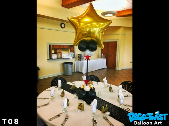 This image shows a decorated dining table with a large gold star-shaped balloon centerpiece. The balloon is anchored by smaller black and white balloons, with a red star accent in the middle. The round table is neatly set with white napkins folded upright in glasses, silverware, and small gold gift boxes at each place setting, creating a formal and celebratory atmosphere.