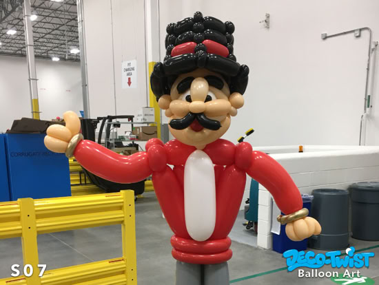 A large balloon sculpture of a circus ringmaster stands indoors. The figure has a black top hat, mustache, and red jacket with outstretched arms, giving a welcoming pose.