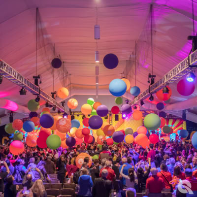 This image captures a lively indoor celebration filled with a large crowd of people. Dozens of oversized balloons in vibrant colors (red, orange, purple, green, and blue) are floating and bouncing above the excited crowd. Many people have their hands raised, reaching for the balloons, while others are taking photos. The atmosphere is festive and energetic, resembling a balloon drop party or grand finale event.